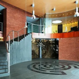 Wnętrze budynku z ceglanymi ścianami, schodami z metalową barierką i dekoracyjnym wzorem labiryntu na podłodze.