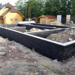 Wylane i zabezpieczone fundamenty pod budowę domu drewnianego, widoczny szalunek i izolacja przeciwwilgociowa, w tle dom w stanie surowym otwartym i zaparkowane samochody.