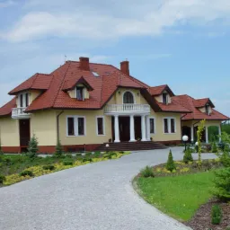 Elegancki, parterowy dom z poddaszem użytkowym, żółtą elewacją, czerwoną dachówką i białymi kolumnami przy wejściu, otoczony zielonym ogrodem z kamienną ścieżką.