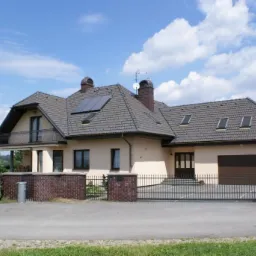 Parterowy dom jednorodzinny z beżową elewacją, szarą dachówką, panelami słonecznymi na dachu i balkonem, otoczony ogrodzeniem z ceglanymi słupkami i metalową bramą.