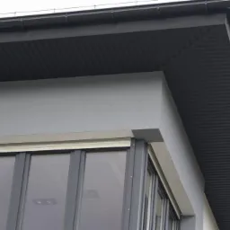 Fragment elewacji budynku z szarą, gładką ścianą, oknem z roletą zewnętrzną i ciemnoszarą podbitką dachową z widocznymi rynnami.