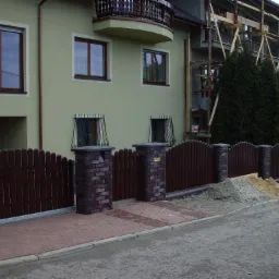 Dwukondygnacyjny dom z zieloną elewacją w trakcie prac wykończeniowych, widoczne rusztowania, brązowe okna, ogrodzenie z drewnianymi sztachetami i ceglanymi słupkami, oraz hałda piasku i żwiru...