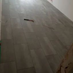 Ułożone szare płytki podłogowe w trakcie prac wykończeniowych, widoczny młotek i poziomica, fragment ściany z widocznymi przewodami.
