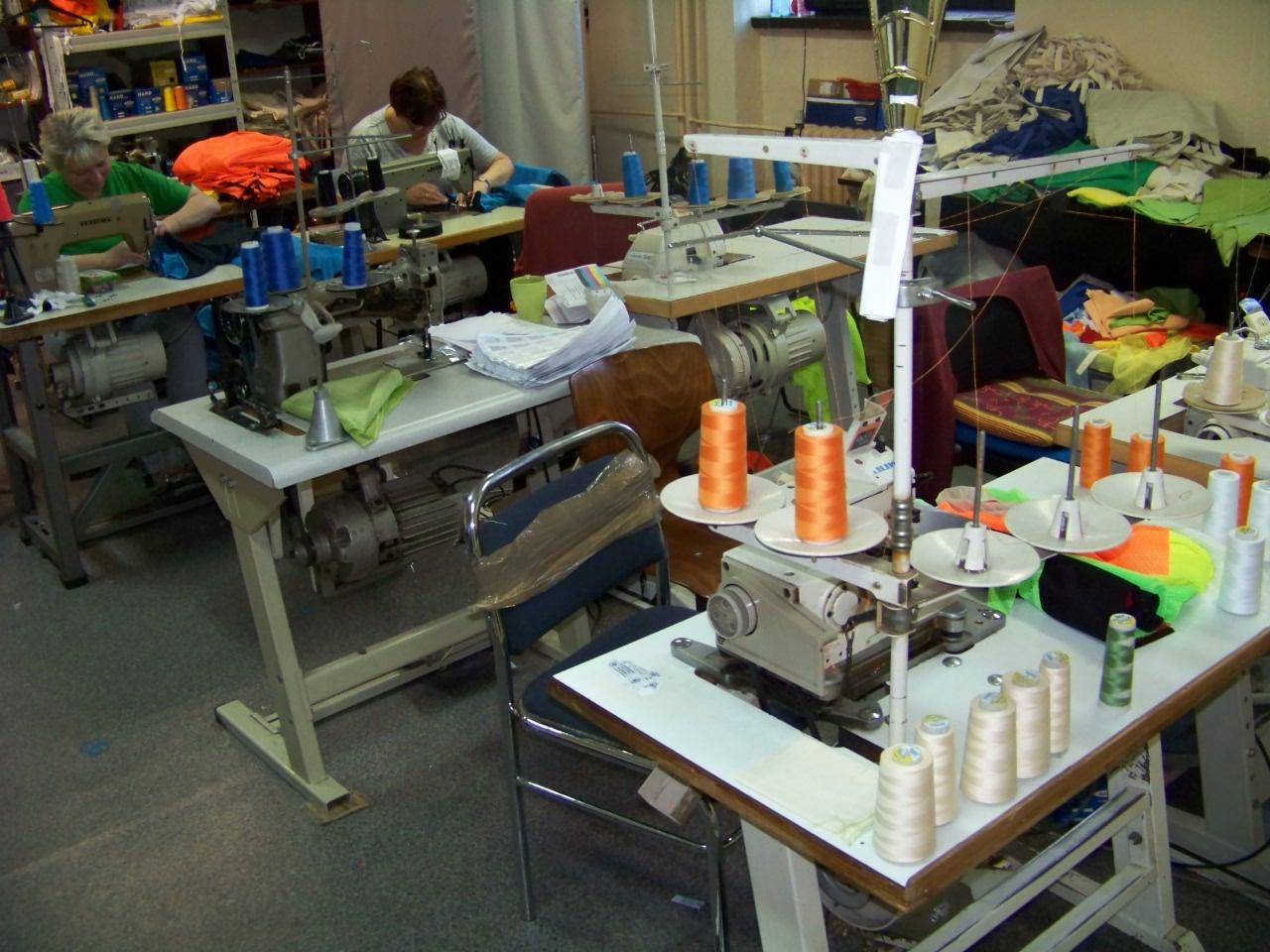 Wnętrze szwalni z maszynami do szycia, nićmi w różnych kolorach i pracującymi osobami. Widoczne detale stanowisk pracy i różnorodność materiałów.