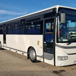 Biały autobus marki Irisbus na kostce brukowej, z widocznymi niebieskimi siedzeniami przez okna. Tablica rejestracyjna widoczna na przodzie pojazdu.