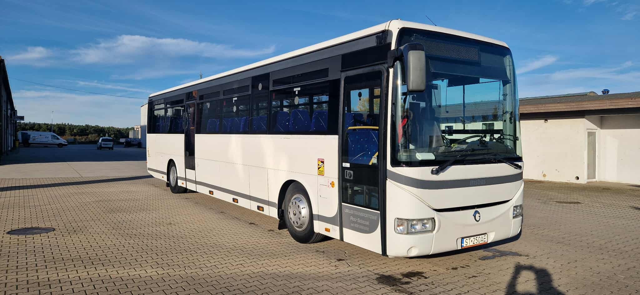 Biały autobus marki Irisbus na kostce brukowej, z widocznymi niebieskimi siedzeniami przez okna. Tablica rejestracyjna widoczna na przodzie pojazdu.