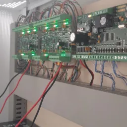 Panel sterowania alarmem z zielonymi diodami LED, widoczne kable i dwa akumulatory ALARMTEC na dolnym planie, zamontowany wewnątrz białej szafki.