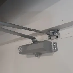 Szary samozamykacz do drzwi zamontowany na białych drzwiach i ścianie, widoczna sprężyna i ramię, zbliżenie na mechanizm.