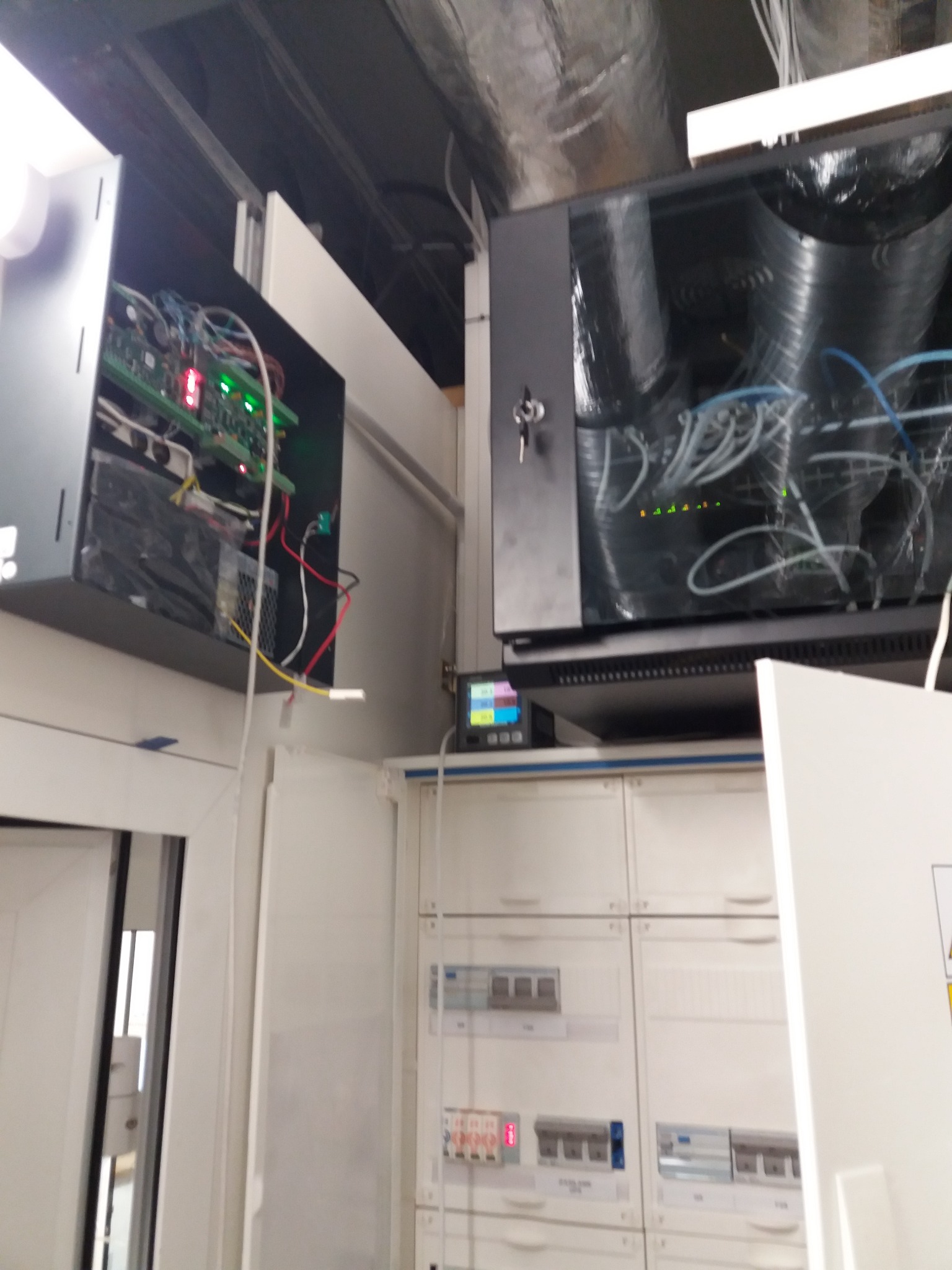 Otwarta szafa telekomunikacyjna z widocznymi kablami i płytą główną z diodami LED, obok skrzynka z bezpiecznikami w pomieszczeniu technicznym.