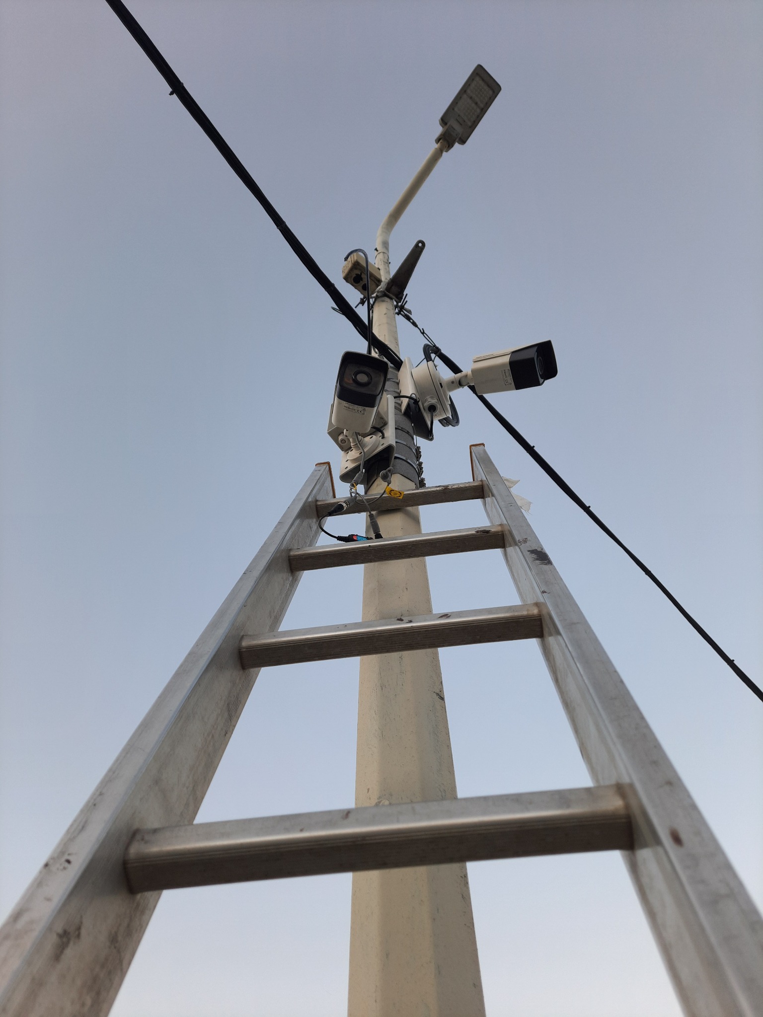 Słup z zamontowanymi kamerami monitoringu, oświetleniem LED i drabina przystawiona do słupa, widok z dołu na błękitne niebo.