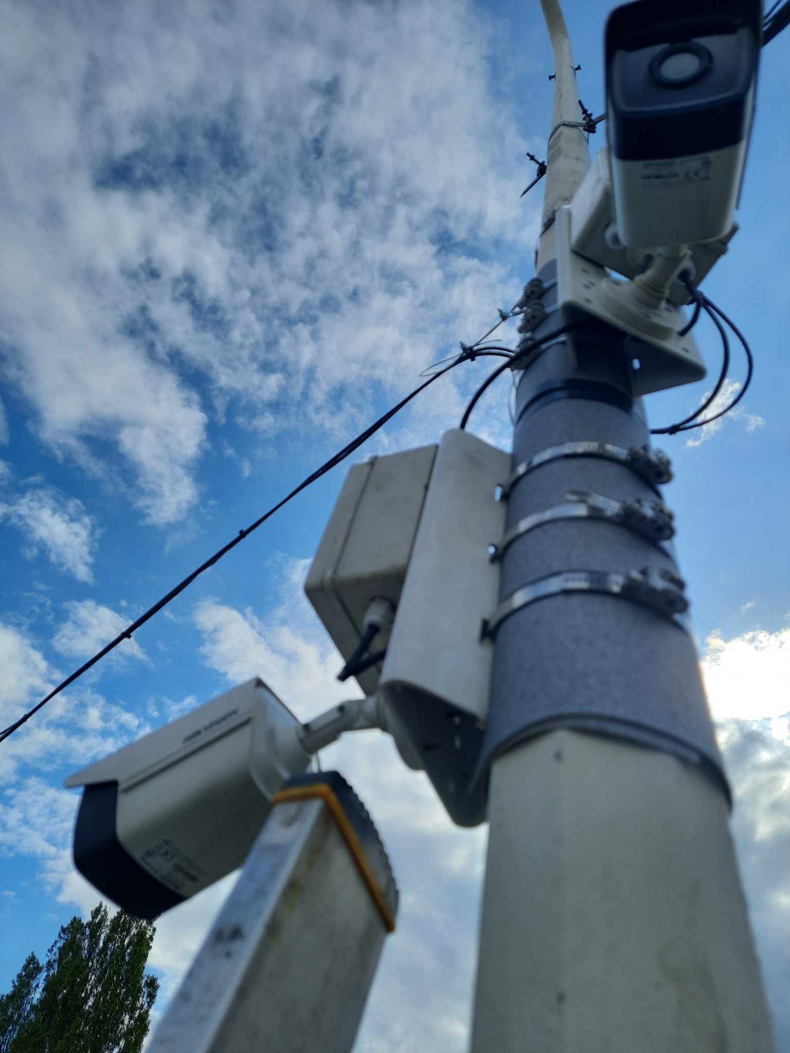 Dwie kamery monitoringu przymocowane do słupa za pomocą metalowych obejm, widok z dołu na błękitne niebo z chmurami, drabina oparta o słup sugeruje montaż lub serwis.
