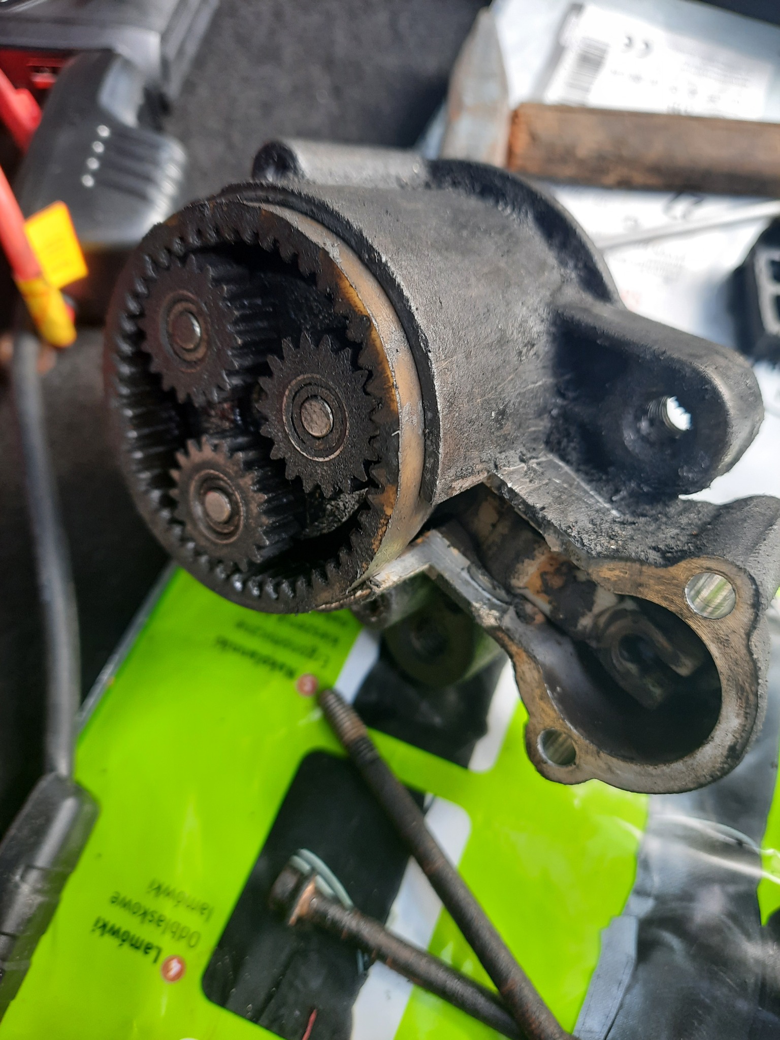 Otwarta obudowa przekładni z trzema zębatkami, widoczne ślady smaru i zużycia, na tle zielonej torby z narzędziami i śrubami.