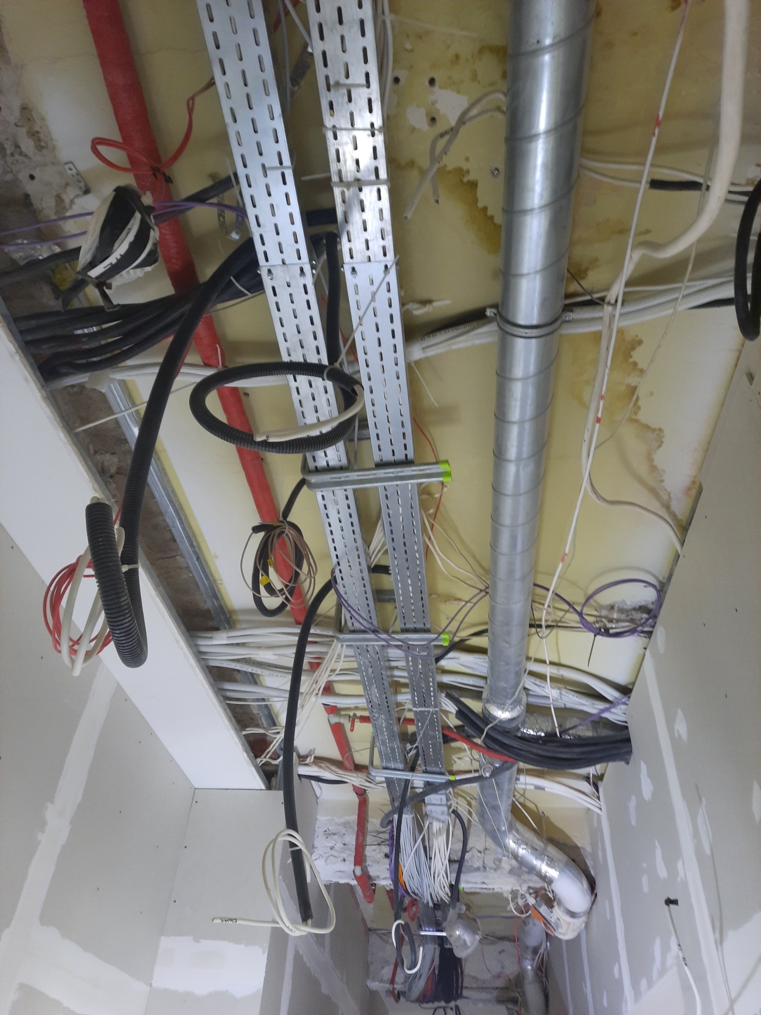 Widok instalacji pod sufitem w trakcie montażu: koryta kablowe, rury wentylacyjne i wiązki przewodów elektrycznych. Widoczne ślady po wierceniu i niedokończone prace wykończeniowe.