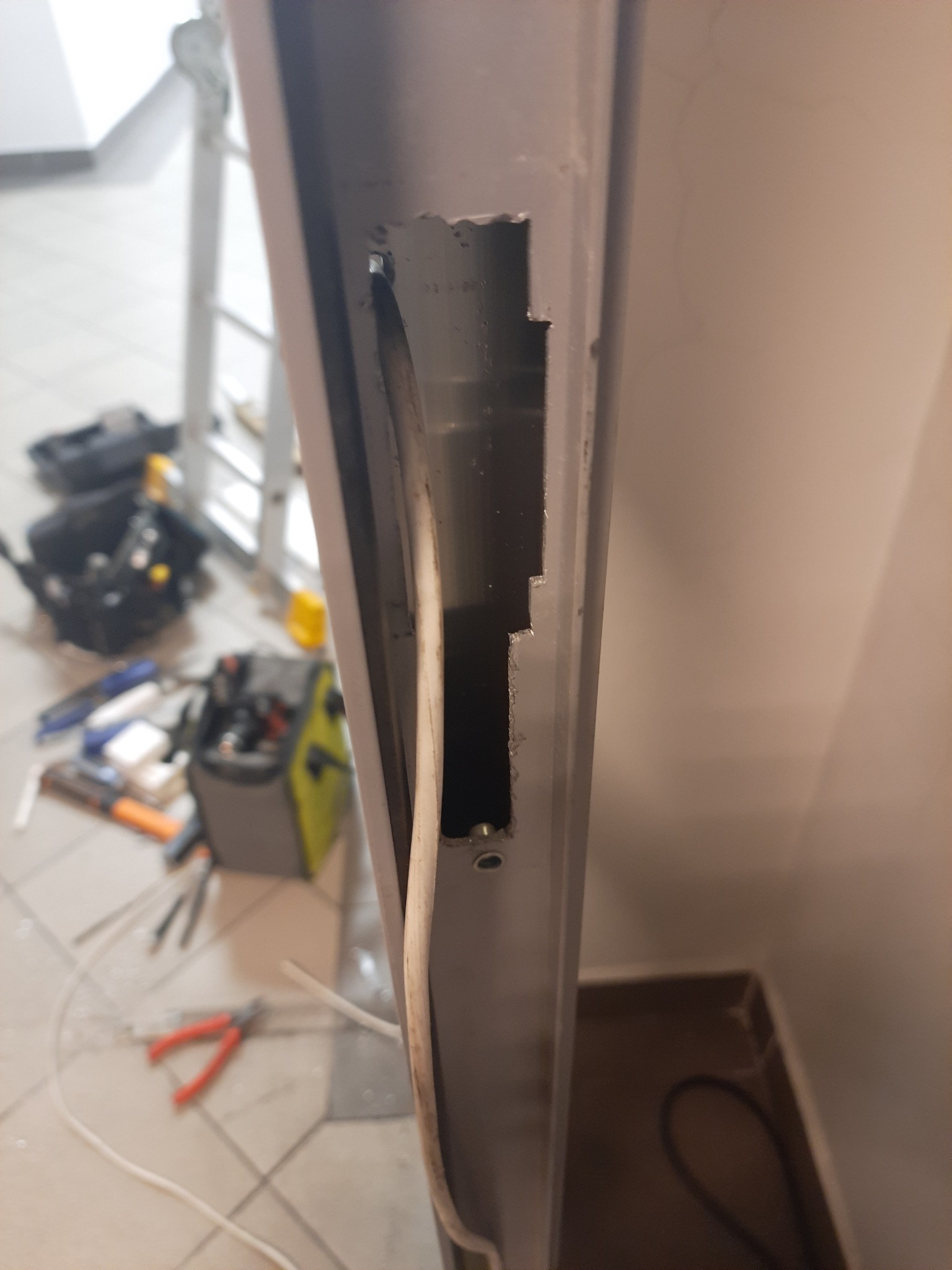 Przygotowanie otworu w metalowej ramie pod przewód, widoczne narzędzia i drabina w tle.