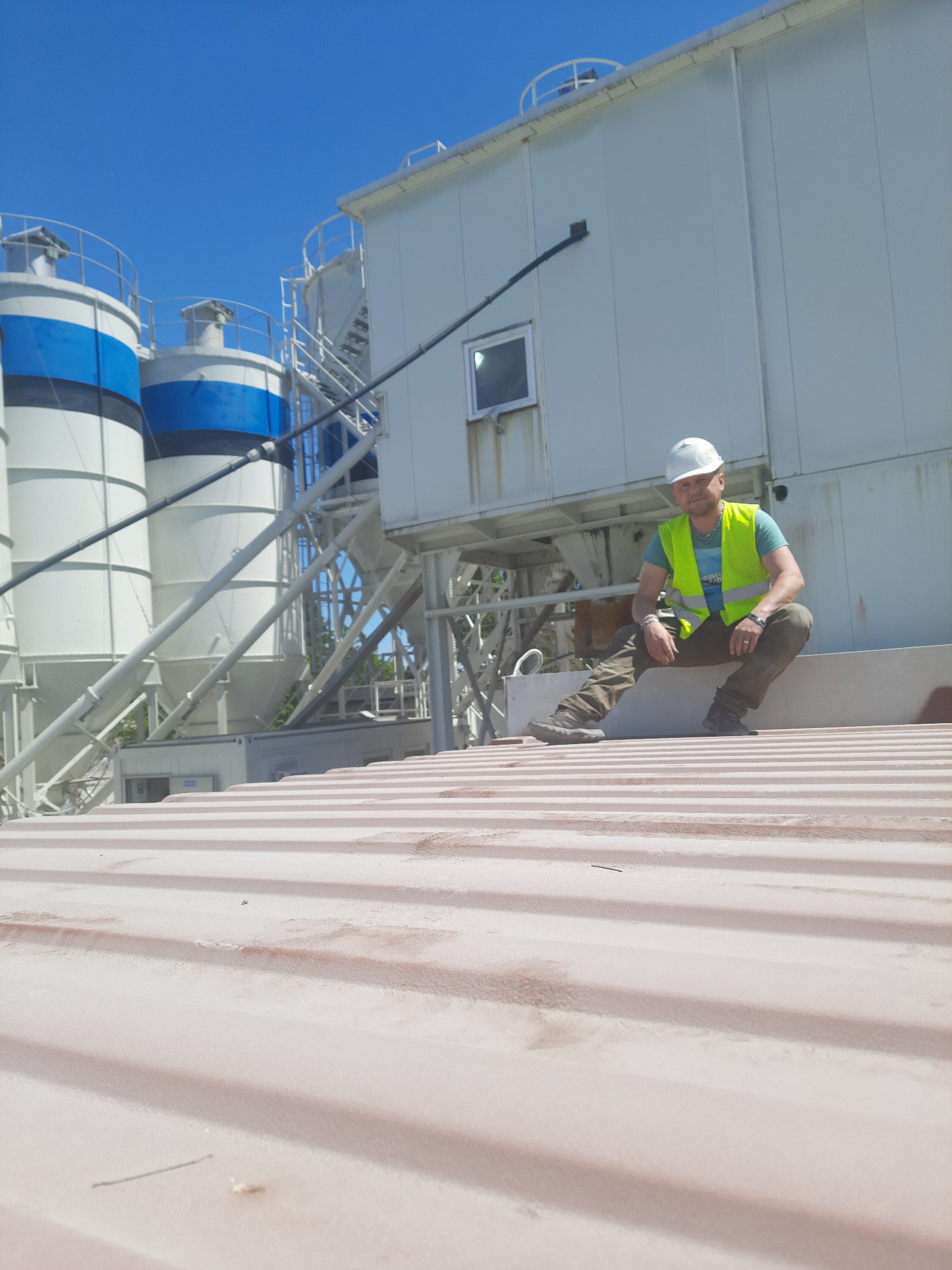 Pracownik w kasku i kamizelce odblaskowej siedzi na dachu przemysłowego budynku z silosami w tle, w słoneczny dzień.