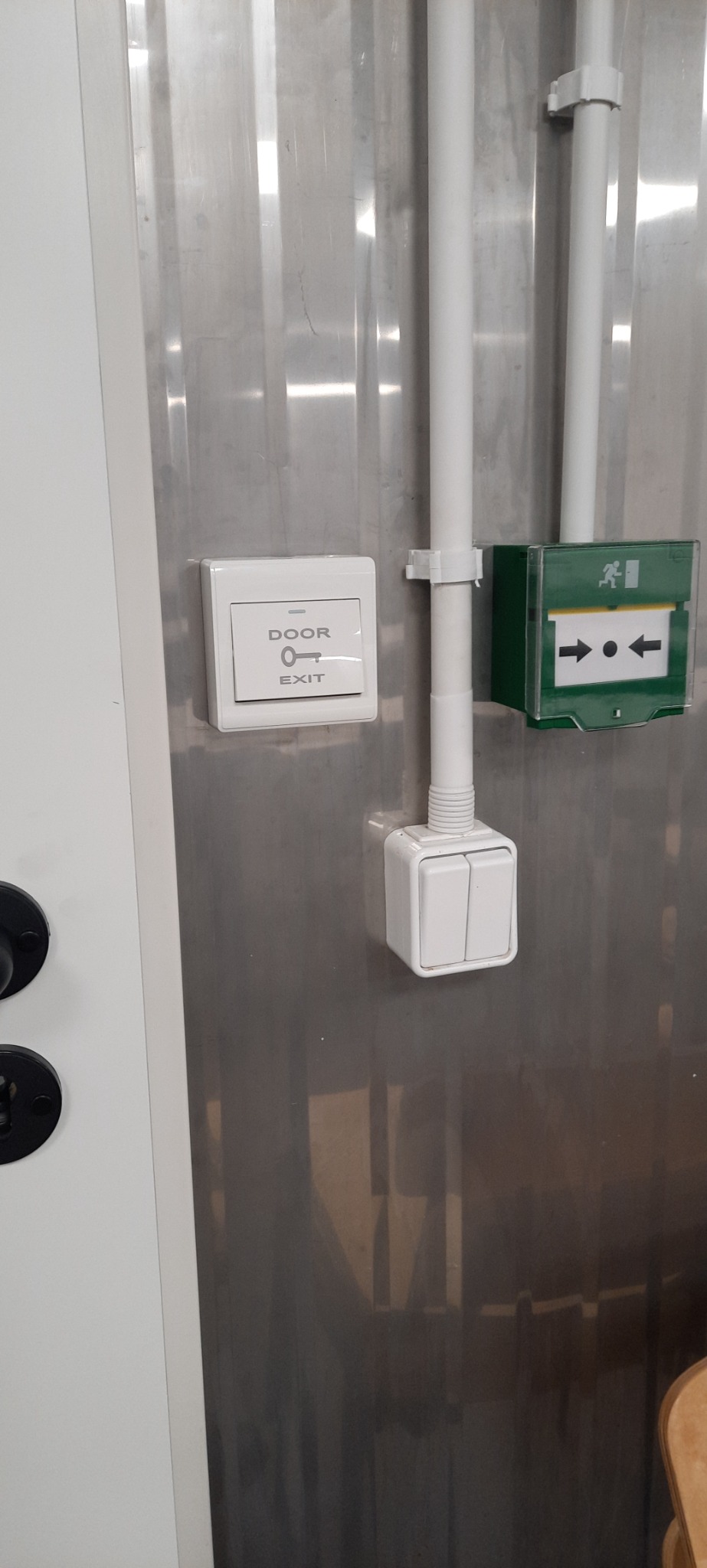 Przycisk otwierania drzwi z symbolem klucza i napisem EXIT, obok zielony znak kierunku wyjścia ewakuacyjnego, białe rury instalacyjne na metalowej ścianie.