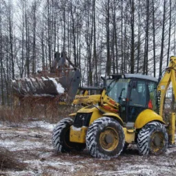 Żółta koparko-ładowarka New Holland z łyżką pełną ziemi i gałęzi na zaśnieżonym terenie, las w tle.
