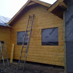 Budowa domu z drewnianych bali, widoczne dwie drabiny opierające się o ściany, okna zabezpieczone folią, fragment dachu z papą, oraz membrana dachowa na ścianie.