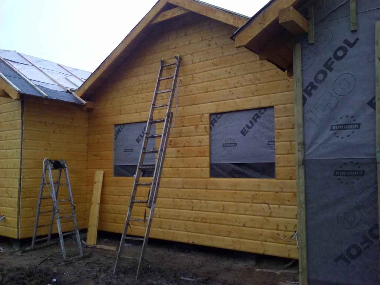 Budowa domu z drewnianych bali, widoczne dwie drabiny opierające się o ściany, okna zabezpieczone folią, fragment dachu z papą, oraz membrana dachowa na ścianie.