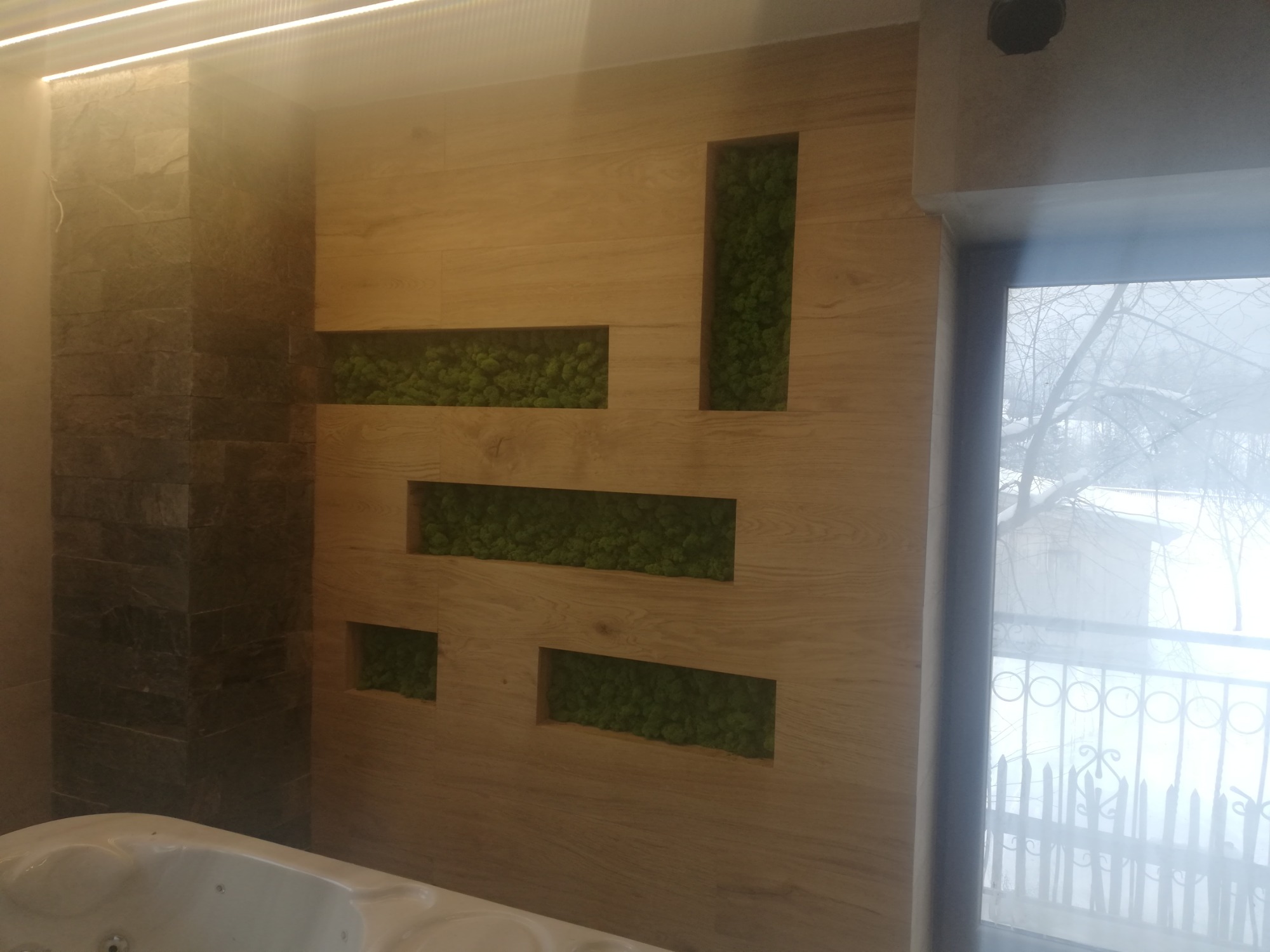 Nowoczesna łazienka z drewnianą ścianą z wnękami wypełnionymi zielonym mchem, obok ściana z kamienia i fragment wanny, za oknem zimowy krajobraz.