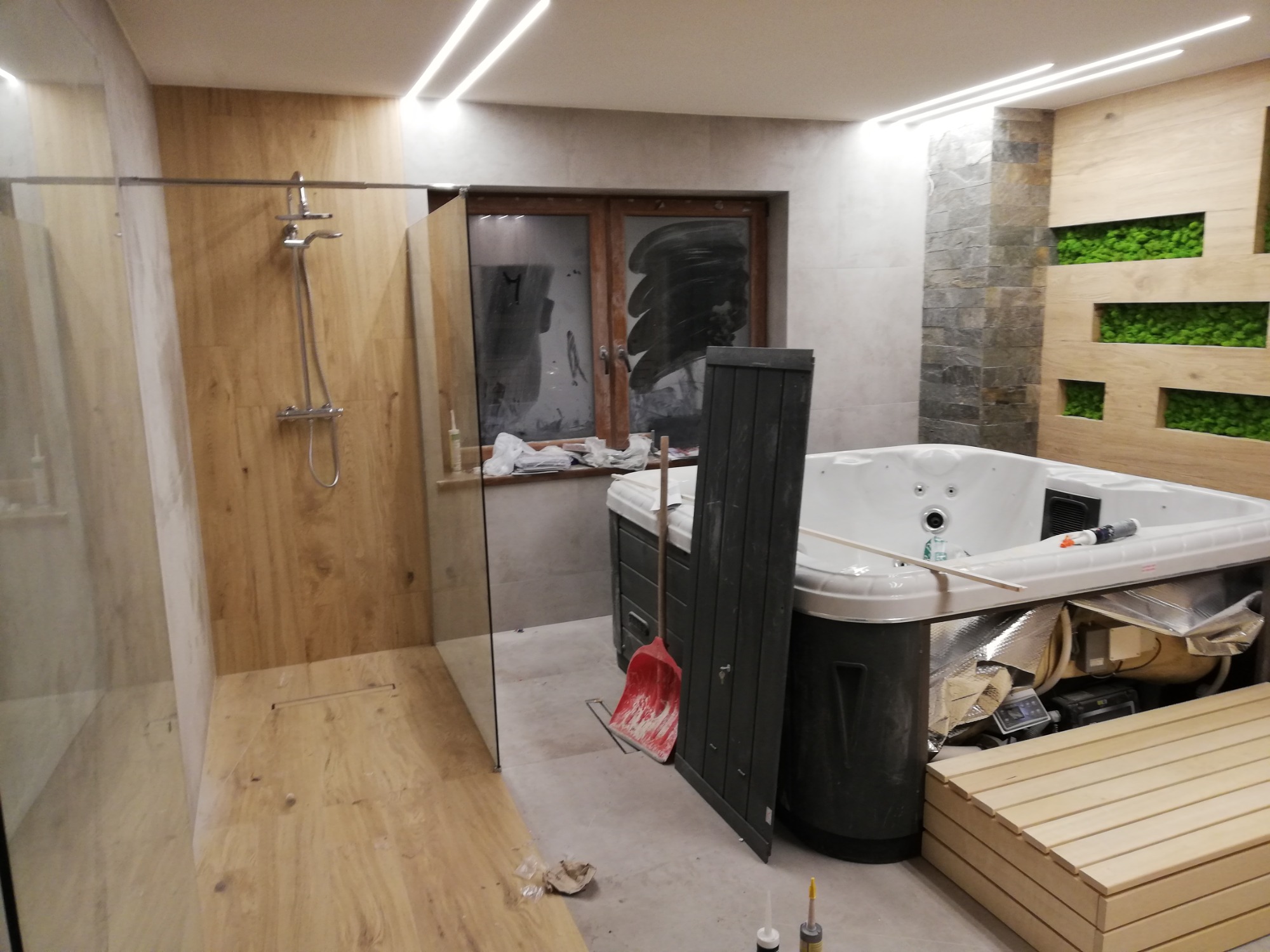 Montaż wanny z hydromasażem i kabiny prysznicowej w łazience, widoczna czerwona szufelka, drewniane panele na ścianie i podłodze, okno zamalowane farbą ochronną.