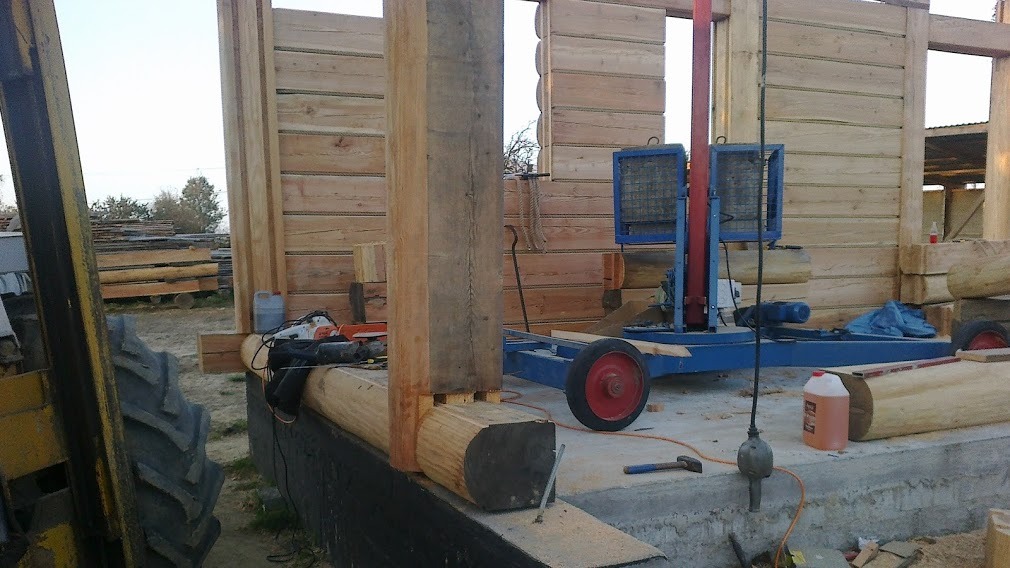 Wznoszenie konstrukcji z drewnianych bali, widoczny fragment ściany, słupy nośne, niebieski podnośnik i narzędzia na placu budowy.