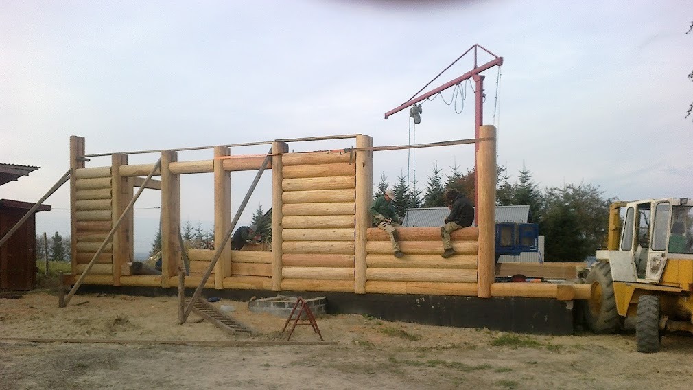 Wznoszenie konstrukcji z bali: widoczny szkielet domu w trakcie budowy, dwóch pracowników siedzi na ścianie, a trzeci pracuje wewnątrz, na zewnątrz stoi żółty traktor z dźwigiem.