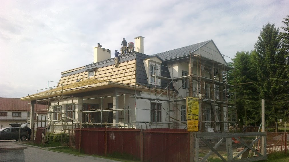 Ekipa dekarzy montuje dachówkę na częściowo wykończonym domu, widoczne rusztowania i przygotowane fragmenty dachu.