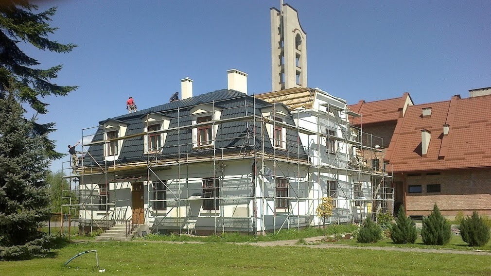 Dwukondygnacyjny dom w trakcie remontu dachu, z czarną dachówką, otoczony rusztowaniami, z pracownikami na dachu, na tle błękitnego nieba i sąsiedniego budynku z czerwonym dachem.