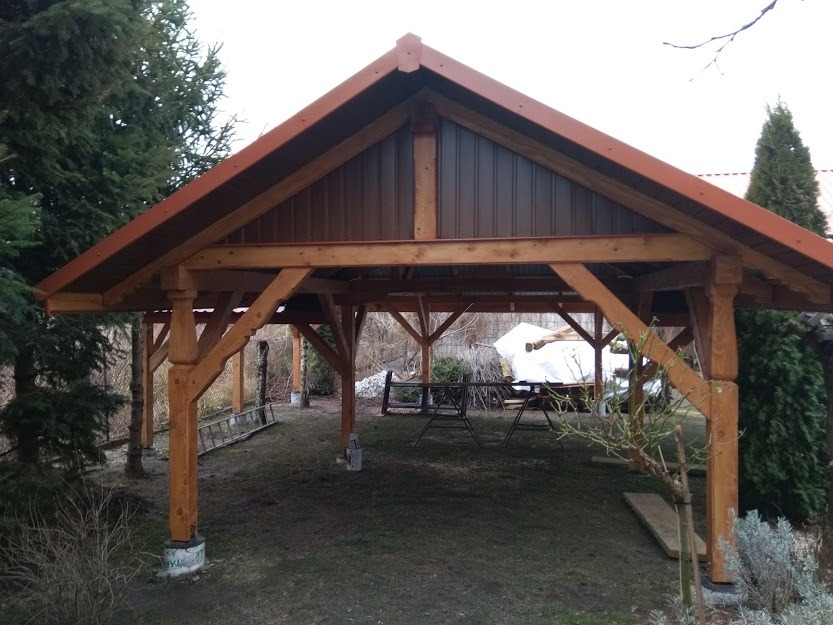 Drewniana wiata z brązowym dachem blaszanym, wsparta na ozdobnych słupach, widoczna w ogrodzie z elementami krajobrazu.