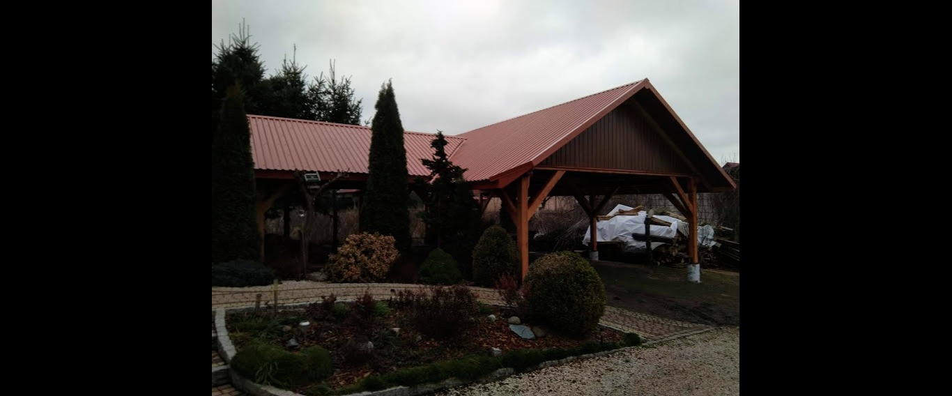 Drewniana wiata z czerwonym, trapezowym dachem, widoczna z zewnątrz, otoczona ozdobnym ogrodem z krzewami i drzewami iglastymi, pod pochmurnym niebem.