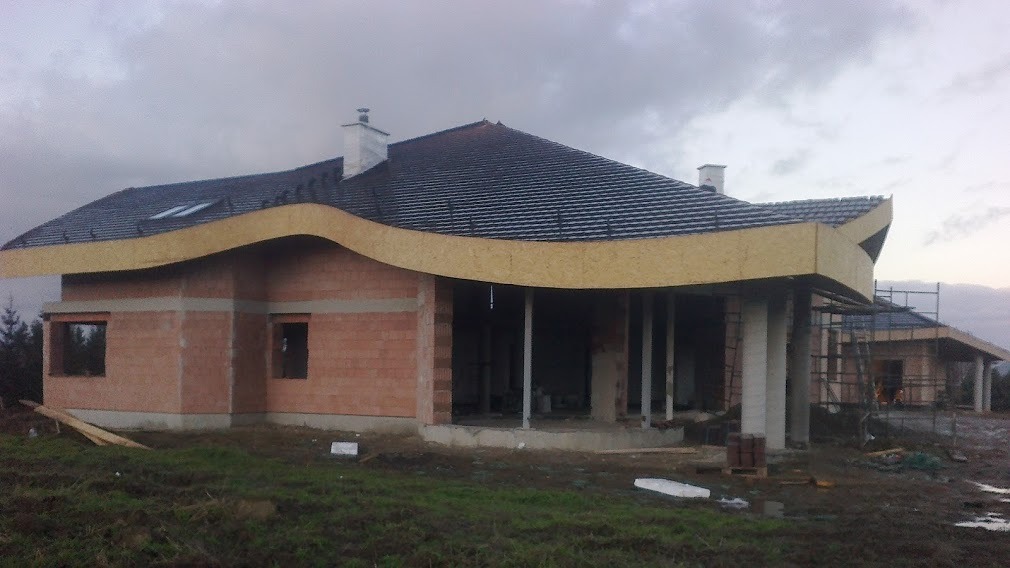 Surowy stan budynku z czerwonej cegły z charakterystycznym, falistym okapem i czarną dachówką, widoczne rusztowania i elementy konstrukcyjne, pochmurne niebo w tle.