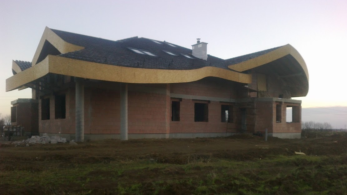 Surowy stan budynku jednorodzinnego z cegły z elementami drewnianymi na dachu i kolumnami, widoczne okna bez szyb i teren budowy.