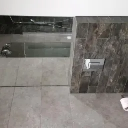 Nowoczesna łazienka z szarą, prostokątną glazurą imitującą kamień na ścianie, podwieszana toaleta z chromowanym przyciskiem spłukiwania i szarą podłogą z dużych płytek, widoczny fragment kabiny...