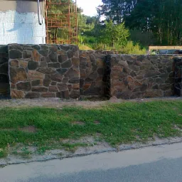 Kamienne murki ogrodowe z nieregularnych, brązowo-szarych kamieni, oddzielone zielonym trawnikiem od asfaltowej drogi. W tle widoczny fragment budynku w trakcie budowy.