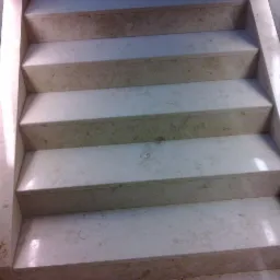 Wykończone schody z jasnego kamienia z widoczną fakturą. Ujęcie z góry podkreśla regularność stopni i precyzję wykonania. Szklana balustrada w tle.