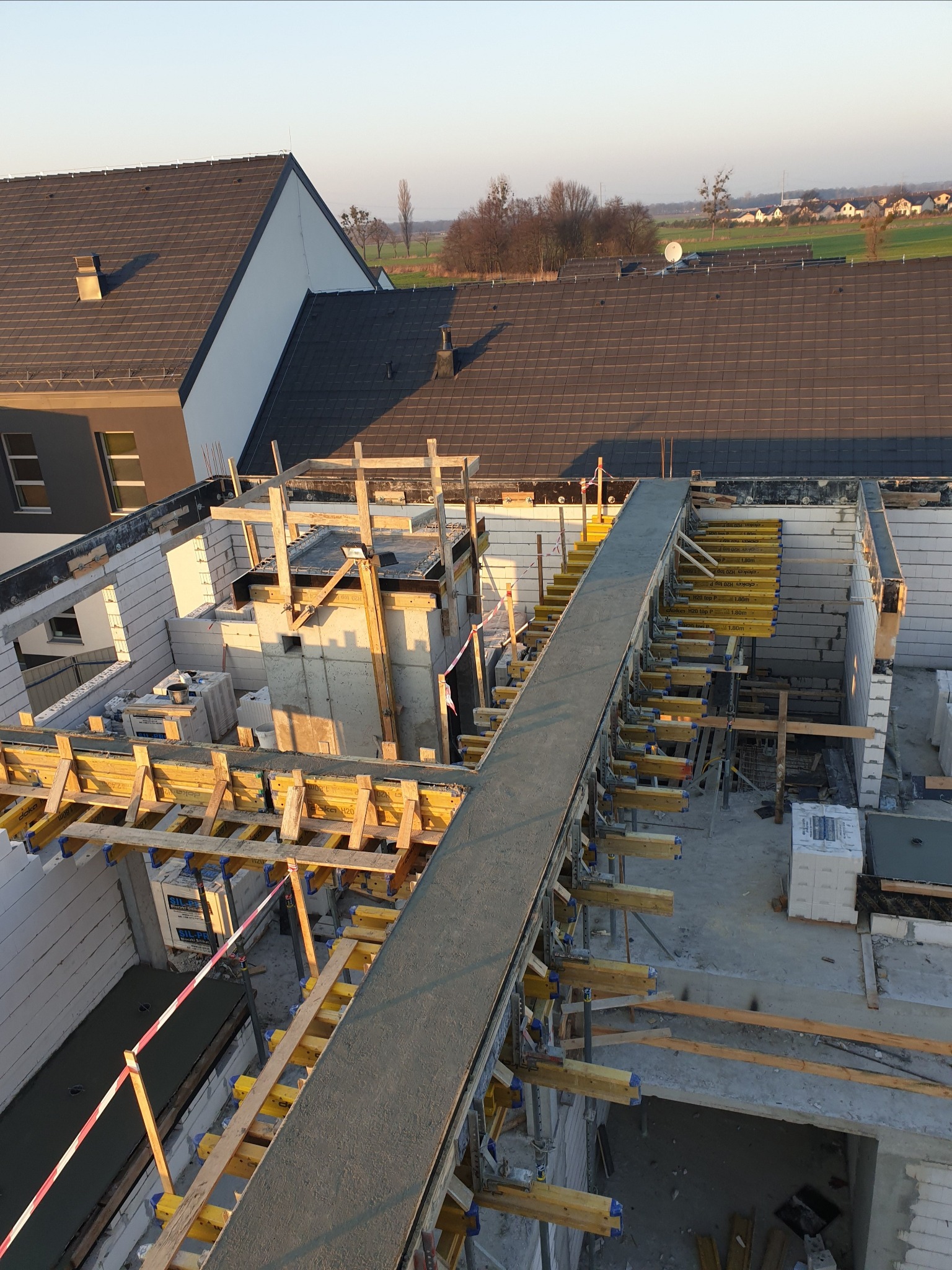 Budowa domu w trakcie wylewania stropu, widoczne szalunki i zbrojenie, w tle domy jednorodzinne.