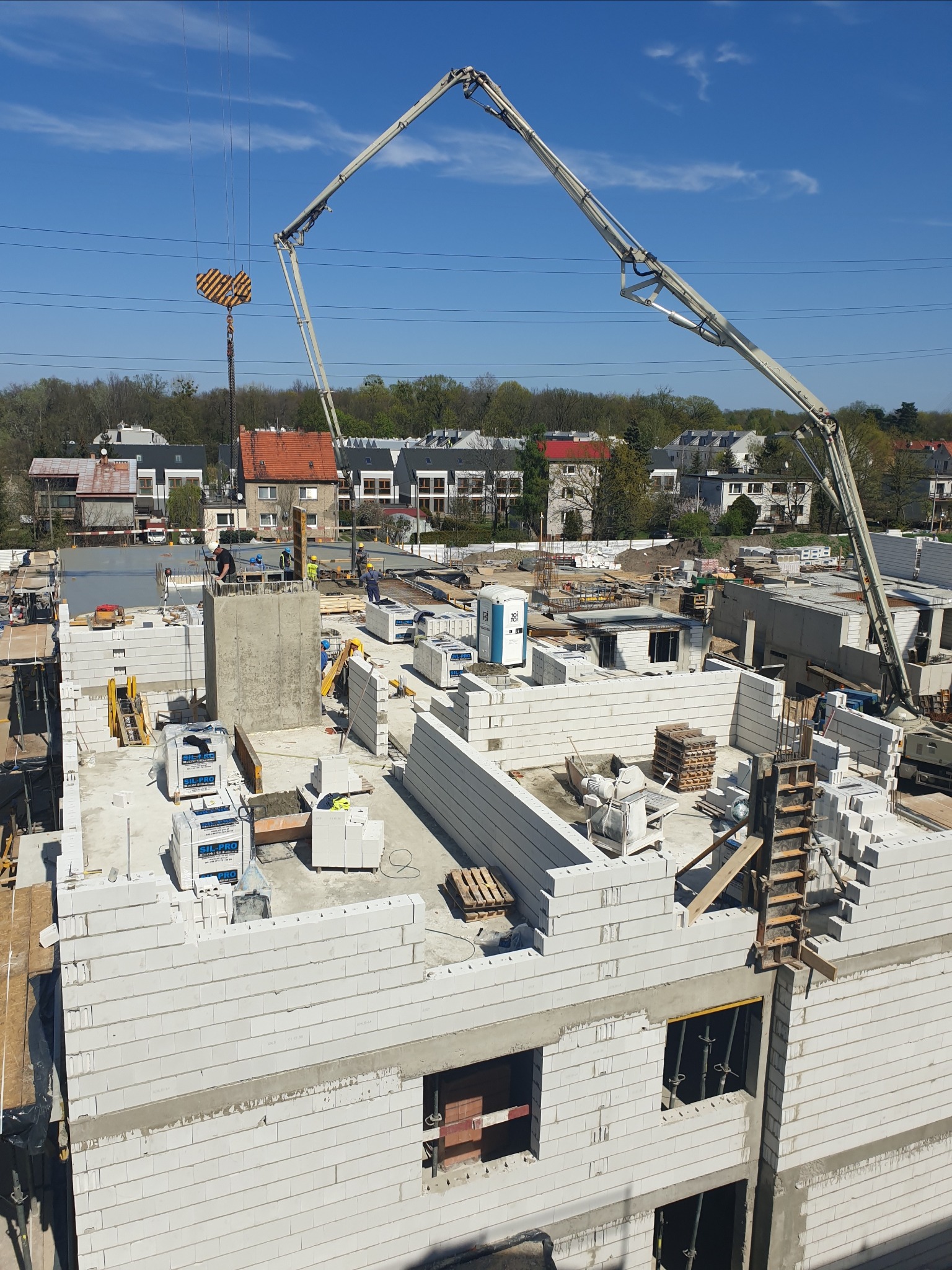 Budowa domu w trakcie wznoszenia ścian z bloczków, widoczny dźwig z pompą do betonu, szalunki, pracownicy w kaskach oraz teren budowy z materiałami i toaletą przenośną w tle.