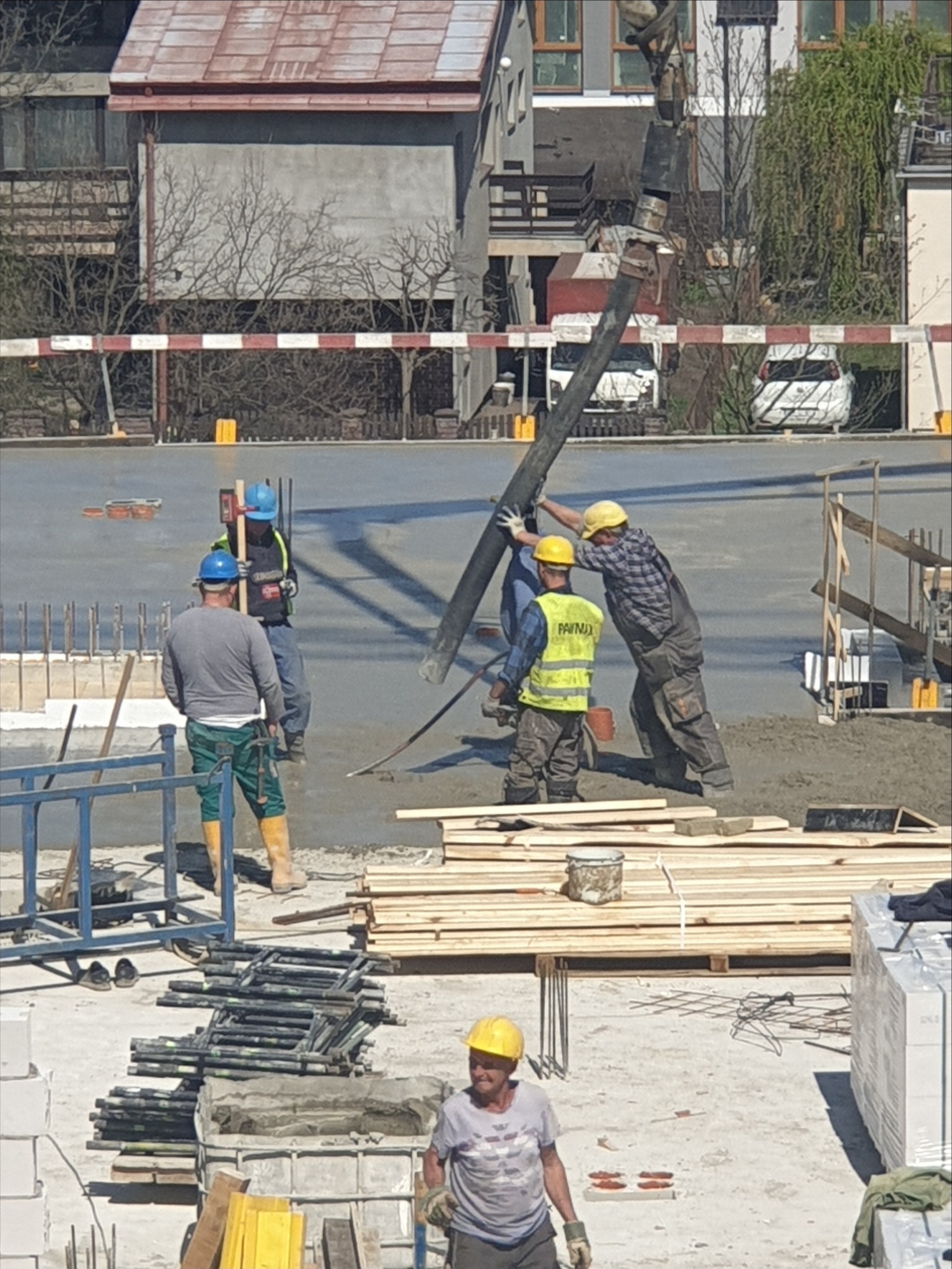 Wylewanie betonu na przygotowany fundament budynku; robotnicy w kaskach kierują wężem pompy betonu, widoczne drewniane szalunki i stalowe elementy zbrojeniowe.