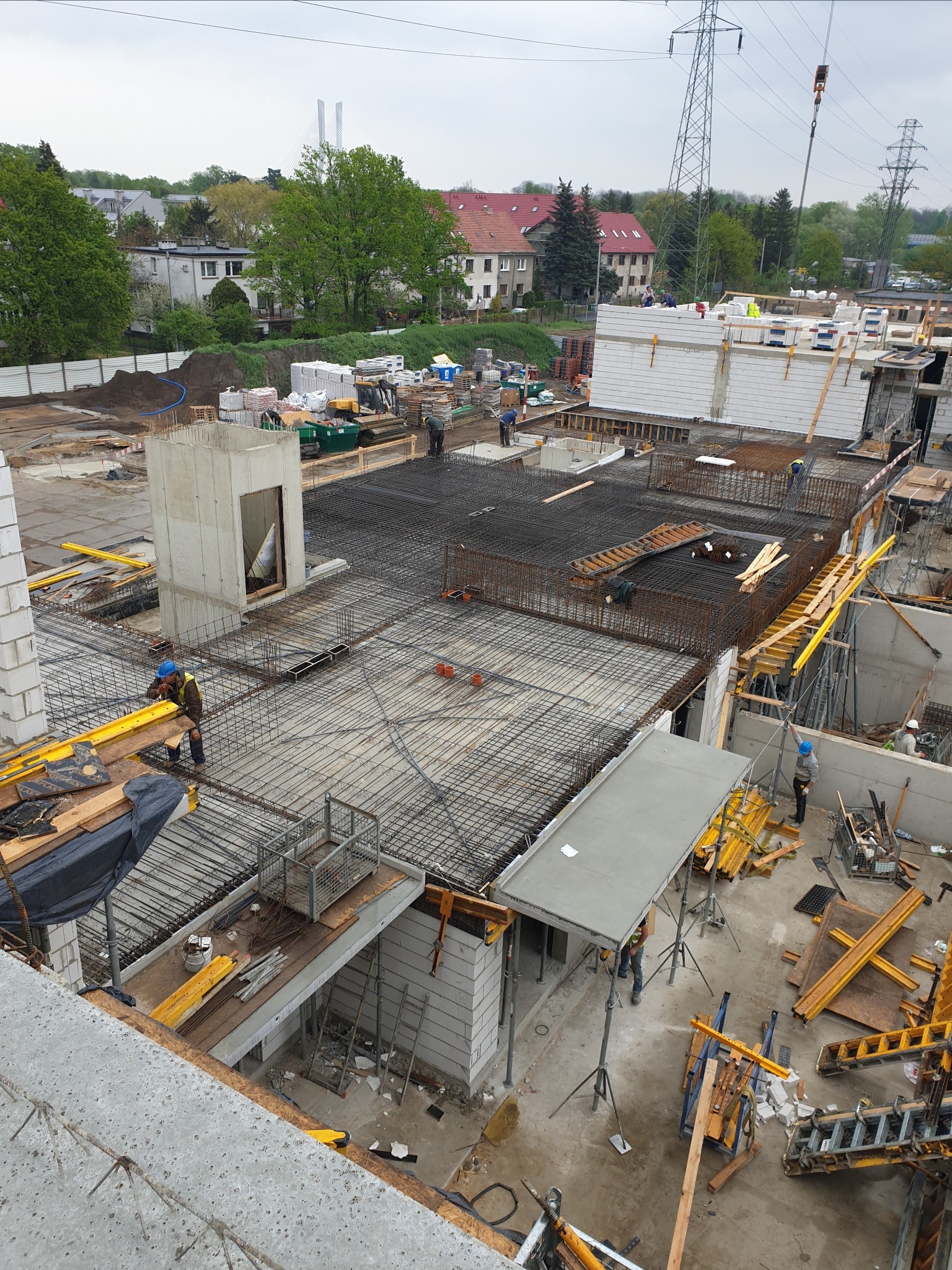 Szeroki kadr z góry na plac budowy z widocznymi zbrojeniami, betonowymi elementami konstrukcyjnymi i pracownikami w kaskach, w tle zabudowa mieszkalna i słupy wysokiego napięcia.