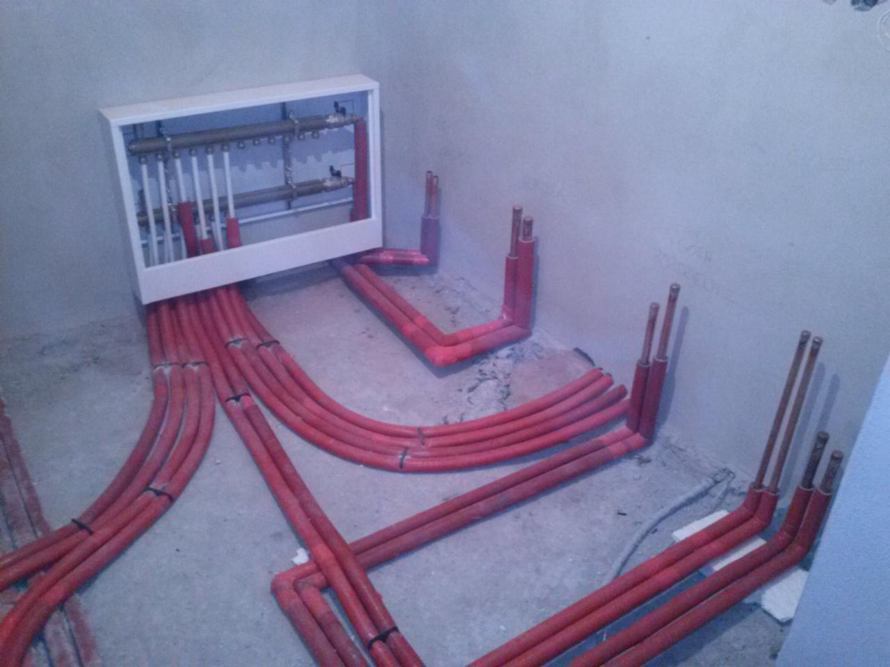 Rozdzielacz centralnego ogrzewania w trakcie montażu: czerwone rury prowadzone w podłodze, widoczne zawory i przyłącza wewnątrz białej skrzynki rozdzielczej na tle surowej ściany.