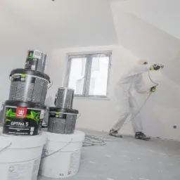 Malowanie natryskowe ścian na biało. Widoczne puszki farby Tikkurila Optiva 5 i pracownik w kombinezonie ochronnym. Wykończenie poddasza.