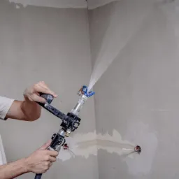 Tynkowanie maszynowe ścian: mężczyzna aplikuje gładź gipsową agregatem, widoczne przewody elektryczne w ścianie, surowe wykończenie wnętrza.