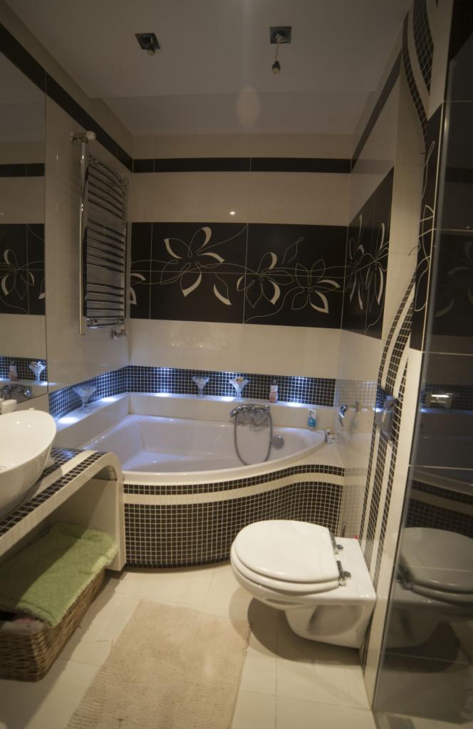 Stylowa łazienka z mozaiką, czarno-białymi płytkami z motywem kwiatowym i podświetlaną wanną. Koszyk z ręcznikami i wisząca toaleta dopełniają aranżację.