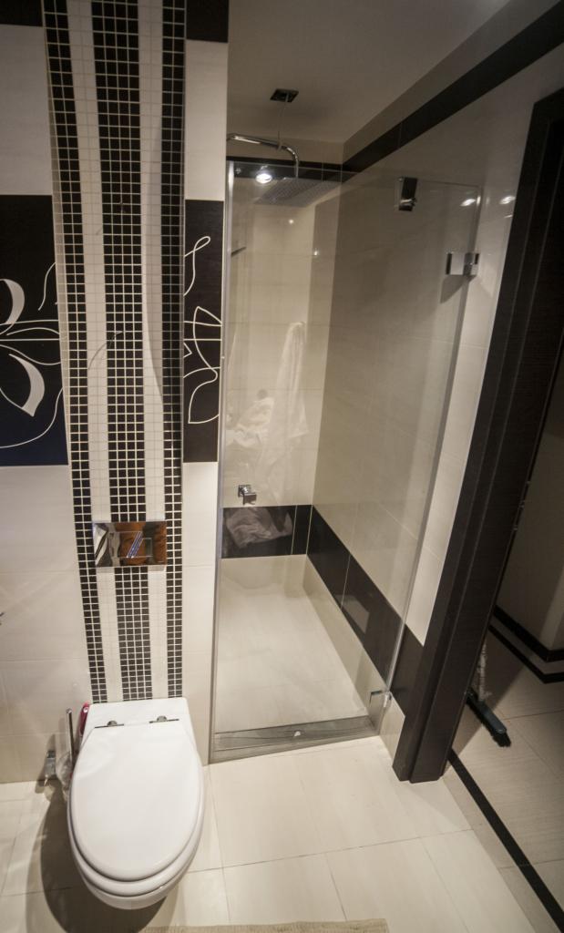 Nowoczesna łazienka z kabiną prysznicową typu walk-in, białą toaletą i dekoracyjnymi czarno-białymi płytkami w pionowe pasy. Minimalistyczny design i eleganckie wykończenie.