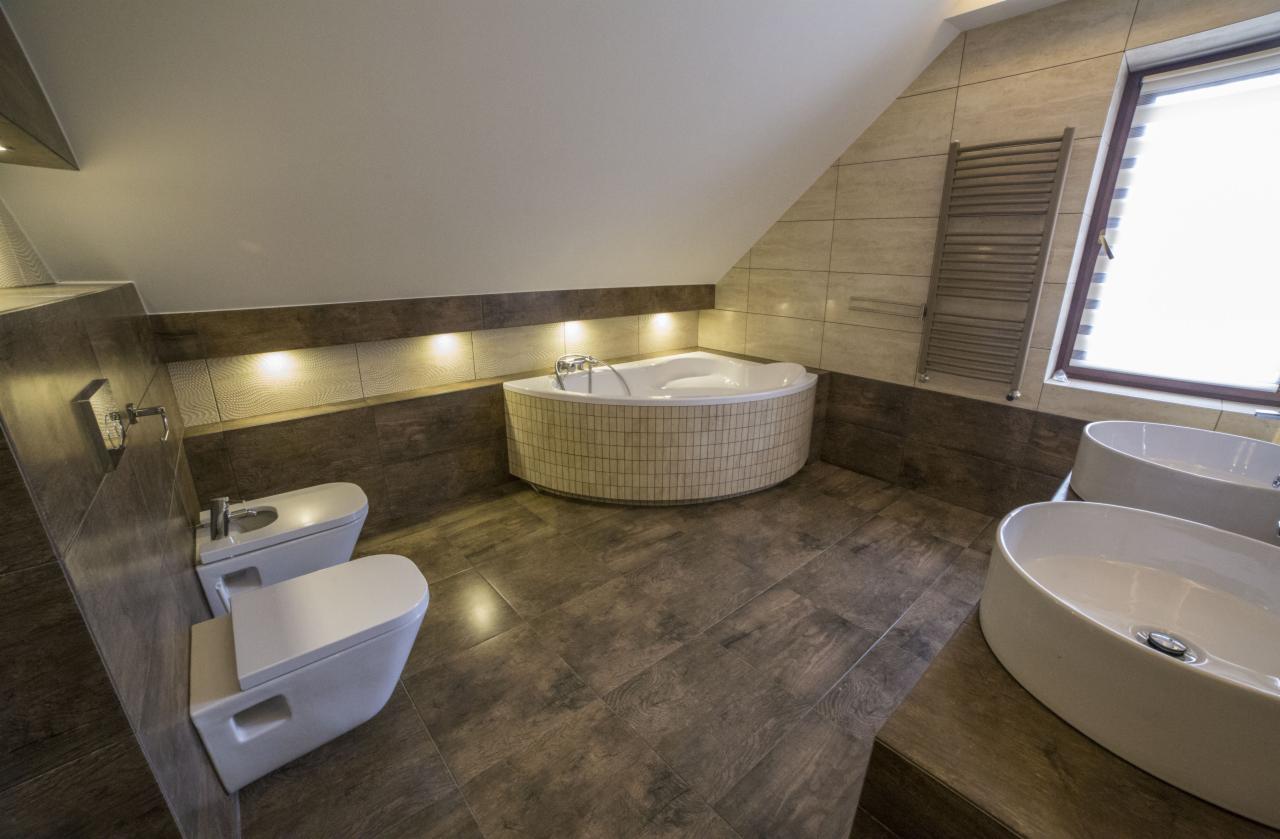 Jasna łazienka na poddaszu z beżowymi płytkami imitującymi drewno, okrągłą wanną w mozaikę, dwiema umywalkami i grzejnikiem drabinkowym przy oknie.