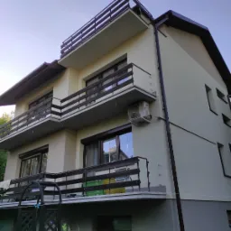 Dwupiętrowy dom z beżową elewacją, brązowymi balkonami i oknami, widoczny z dołu pod kątem, z klimatyzatorem na ścianie.