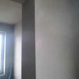 Świeżo otynkowana ściana wewnętrzna w odcieniach szarości, widoczny fragment okna i narożnik ściany z wyraźnym załamaniem światła.