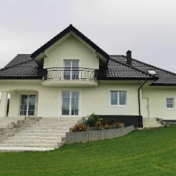 Jasnożółty dom jednorodzinny z czarnym dachem z dachówki ceramicznej, białymi oknami i balkonem z kutą balustradą, widok z dołu na elewację frontową i schody wejściowe.