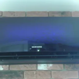 Czarny klimatyzator Samsung zamontowany na ścianie z cegły, wyświetlający temperaturę 24 stopnie Celsjusza, widoczne odbicie w błyszczącej obudowie.